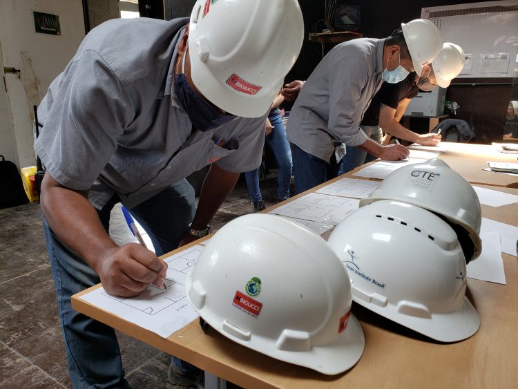Colaboradores MBigucci preenchem texto ao longo do treinamento Lean Construction (construção enxuta). Capacetes são vistos à frente.